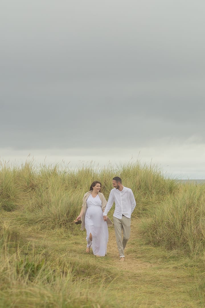 A couple walking through the dunes holding hands. Pregnancy photography