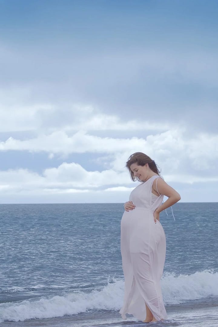 A pregnant woman stood by the sea with her hand on her stomach